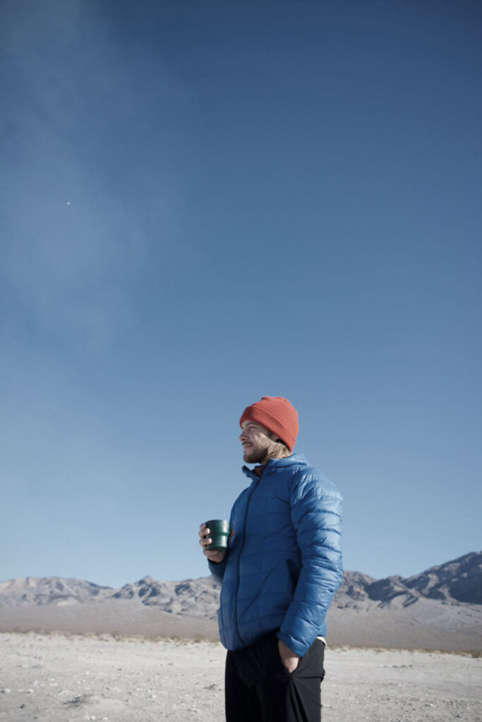 Guy holding a cup of coffee in a puffy jacket and toque, with a blue sky in the background.